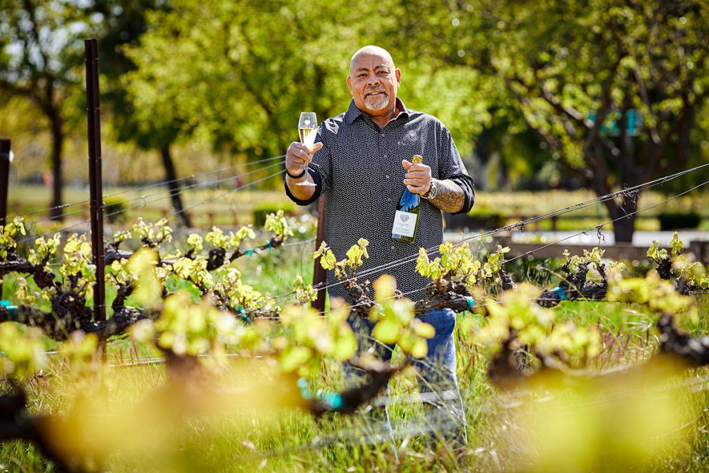 Phil Long of Longevity Wines, standing in the vineyard on a sunny day holding out a glass of sparkling white wine.